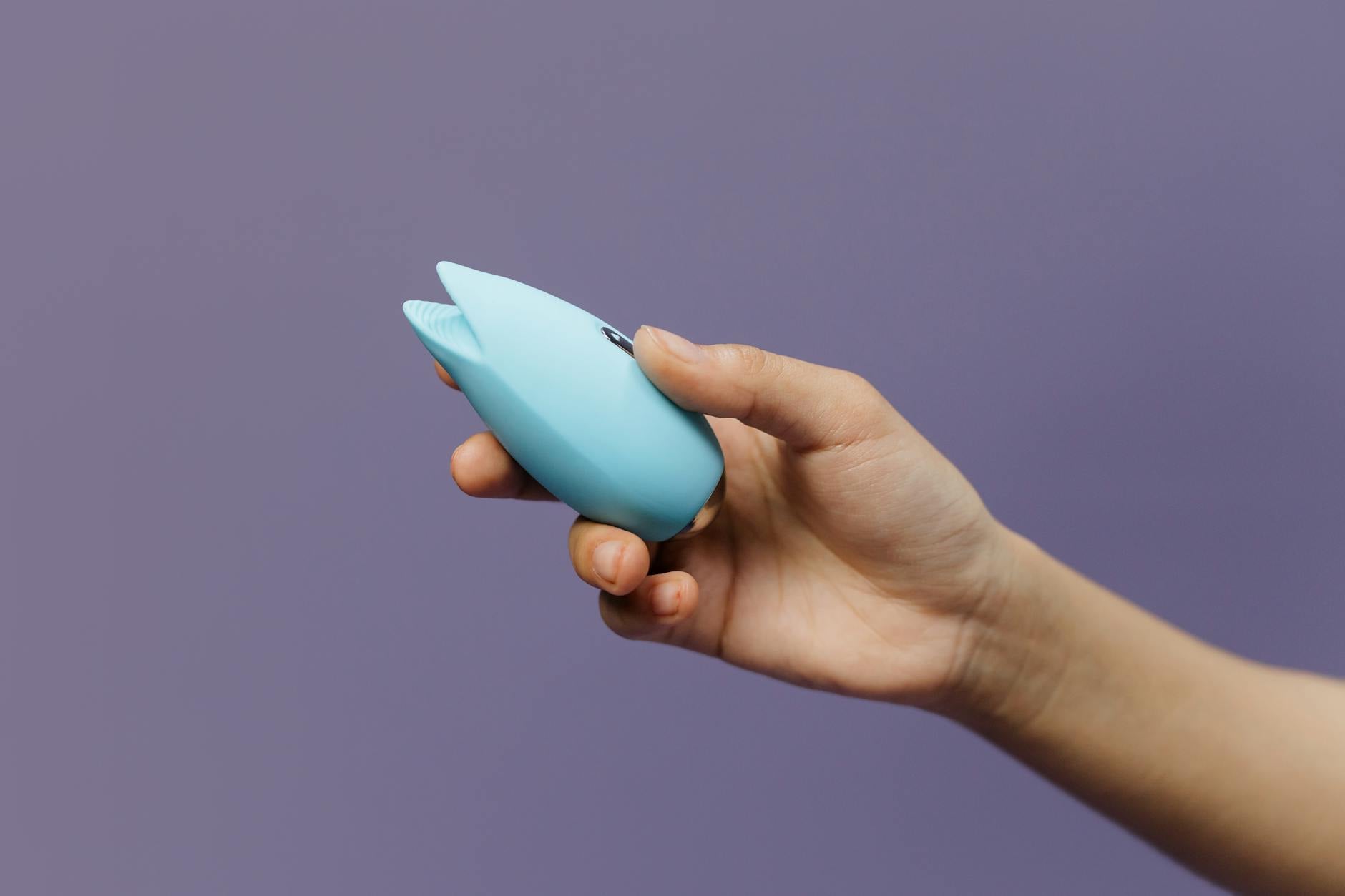 A blue silicone clitoral vibrator held in hand against a purple background, demonstrating modern pleasure tools